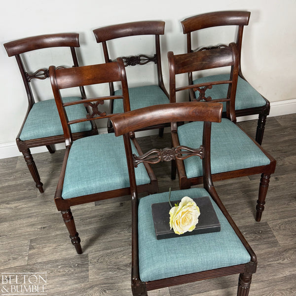 Six vintage mahogany dining chairs arranged in a staggered layout on wooden flooring, featuring teal linen upholstered seats and carved back details, with a single chair in front displaying a closed book topped with a pale yellow rose.