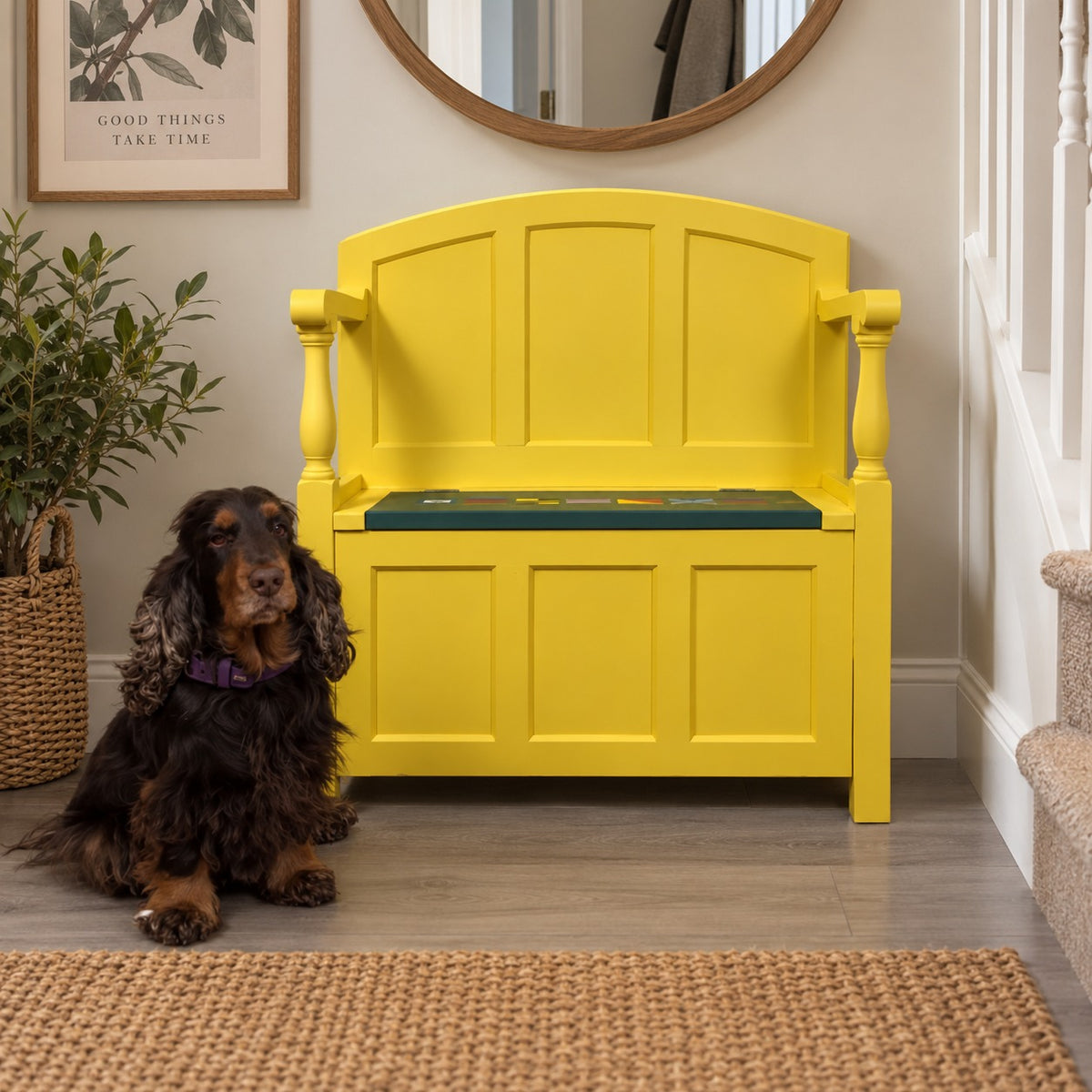 A bright yellow wooden bench with a storage compartment underneath sits in a home entryway next to a black and tan Cocker Spaniel dog. The bench has a dark green cushion on the seat.