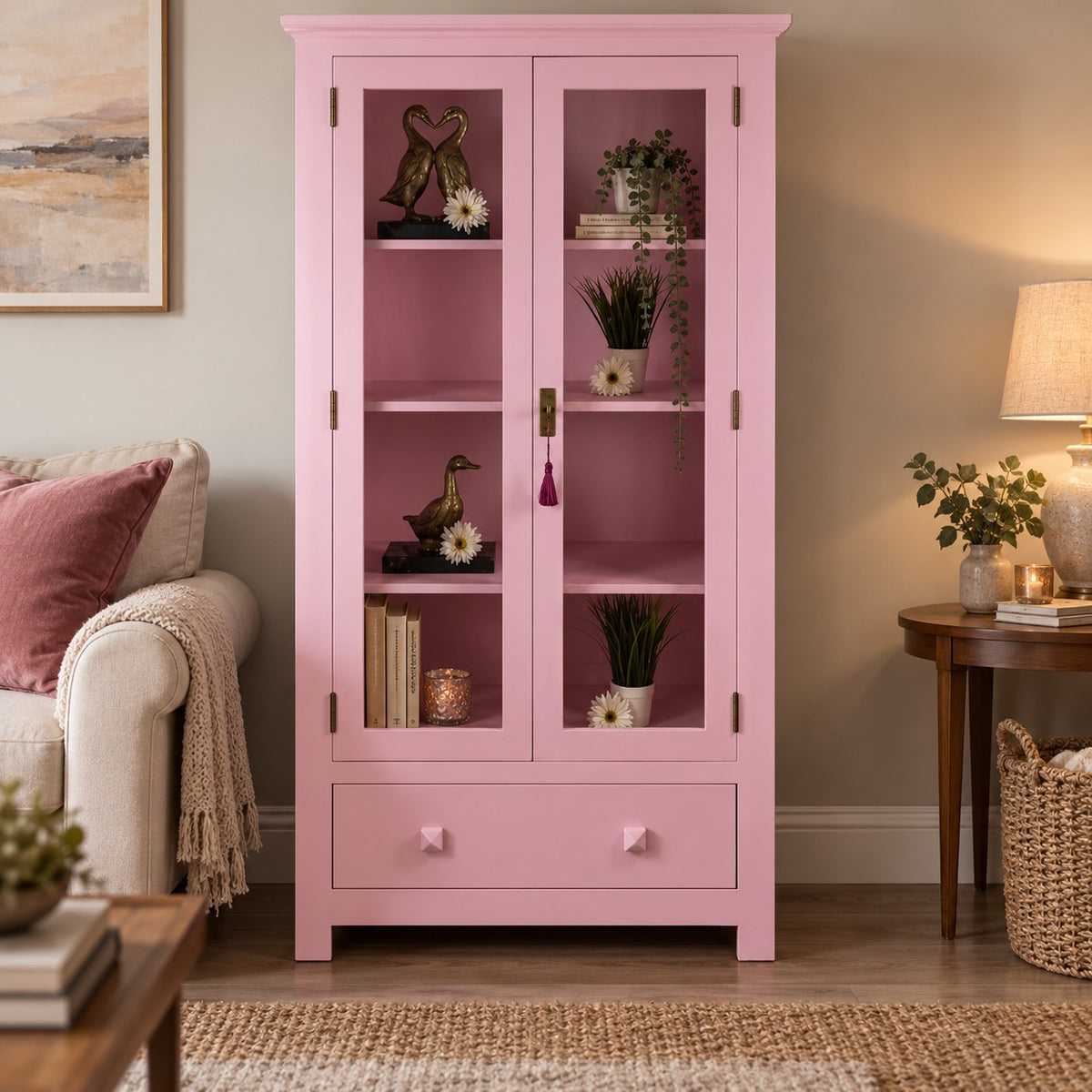 A pink wooden display cabinet with two glass-paneled doors and a single drawer at the bottom, styled in a cozy living room setting next to a sofa and side table.